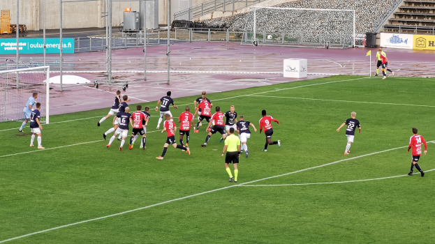 Gruppe von Menschen, die Fußball auf einem Feld mit Zuschauern in Stadionrängen, umgeben von Pfählen, Schildern mit Text, Geländern, Stufen und anderen Gegenständen spielen.