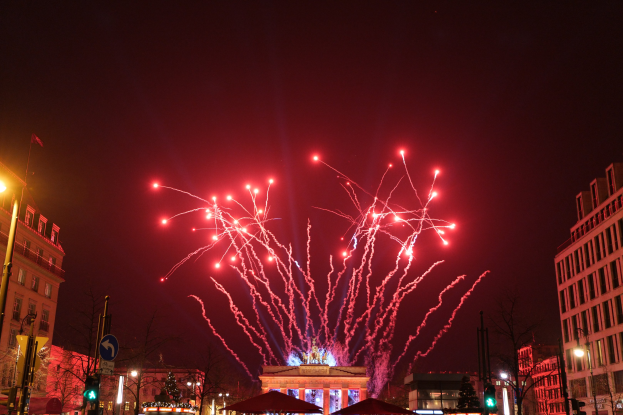 Eine Stadtstraße an Silvester in Berlin, mit Gebäuden, Bäumen, Laternen, Ampeln, Schildern, Zelten, Menschen und einem Feuerwerk erleuchteten Himmel.