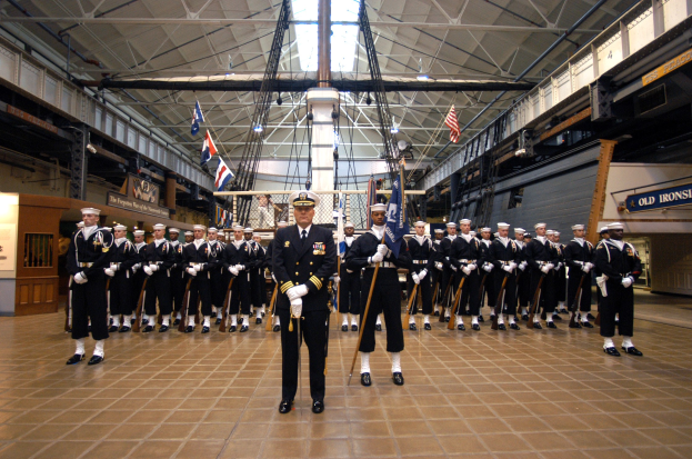 Eine Gruppe uniformierter Männer mit Mützen und Gewehren steht in Formation mit Fahnen und Wandtafeln in einem großen Raum.