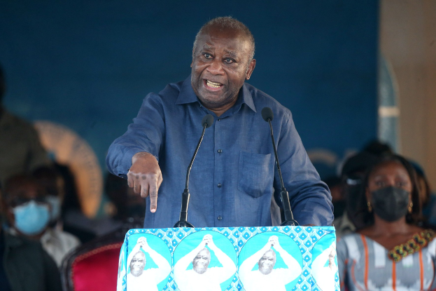 Ein Mann in einem blauen Hemd spricht an einem Pult mit zwei Mikrofonen vor einem sitzenden Publikum, das Masken trägt, und einer Wand im Hintergrund.