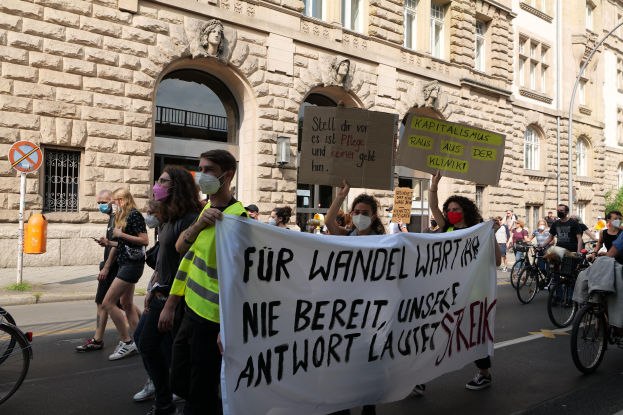 Eine Gruppe von Menschen marschiert auf einer Demonstration durch eine Stadtstraße, einige halten Schilder und andere fahren Fahrräder, im Hintergrund ist ein historisches Gebäude mit Rundbogenfenstern, Säulen und Skulpturen zu sehen.