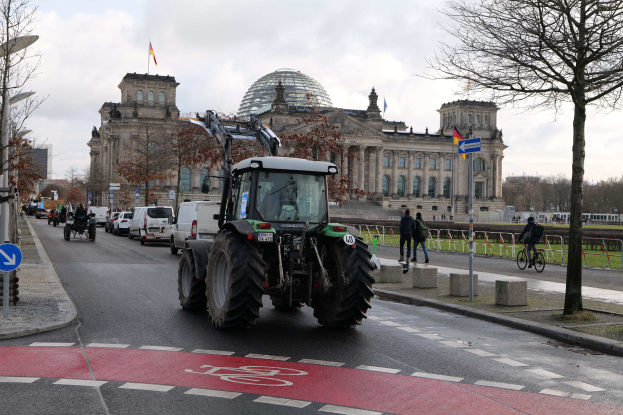 Traktor fährt eine Straße entlang, die von Bäumen und Laternen gesäumt ist und am Reichstaggebäude in Berlin, Deutschland, vorbeifährt, mit Fußgängern und Radfahrern auf dem Gehweg und Fahnen, die das Gebäude schmücken.
