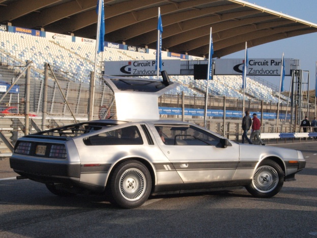 Ein silberner DeLorean DMC-12 fährt auf einer Straße, umgeben von Menschen, Fahnen, Schildern, Geländern, Stühlen und einer Hütte, mit Bäumen und einem klaren blauen Himmel im Hintergrund.