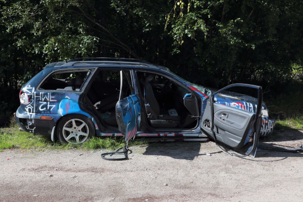 Ein beschädigtes Auto am Straßenrand mit Gras und Bäumen im Hintergrund.