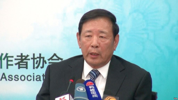 Ein Mann im Anzug und Krawatte sitzt an einem Tisch mit Mikrofonen und hält eine Rede mit einem Banner im Hintergrund.