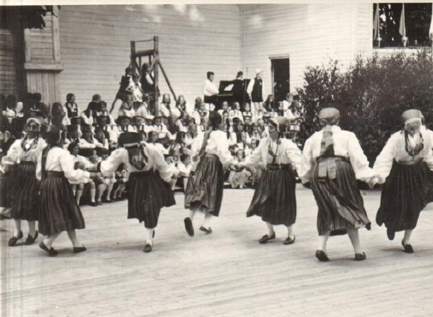Eine Gruppe von Frauen, die vor einem gemischten Publikum aus sitzenden und stehenden Menschen tanzt, mit einem Gebäude, Pflanzen und einem hölzernen Objekt im Hintergrund, dargestellt in Schwarz-Weiß.