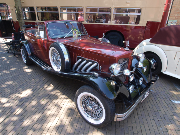 Ein Vintage-Auto neben einem Doppeldeckerbus auf einem Kopfsteinpflasterweg geparkt, mit Stühlen und einem Baum im Vordergrund, einem Gebäude mit Fenstern im Hintergrund und Menschen im Bus zu sehen.