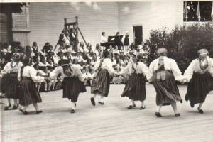 Eine Gruppe von Frauen tanzt vor einem gemischten Publikum aus sitzenden und stehenden Menschen, mit einem Gebäude, Pflanzen und einem hölzernen Objekt im Hintergrund, dargestellt in Schwarz-Weiß.