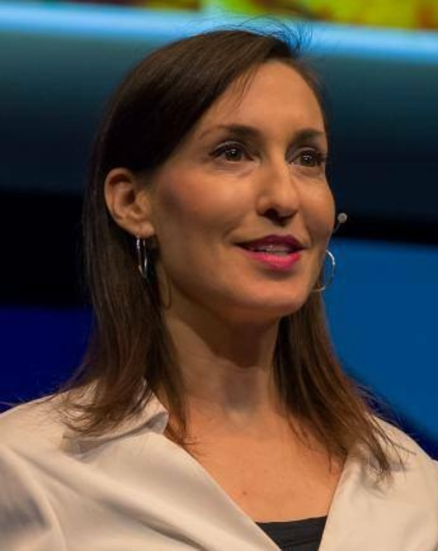 Frau in weißer Jacke und schwarzem Oberteil vor einem blauen und gelben Hintergrund sitzend, trägt Ohrringe und ein Mikrofon während eines Interviews.