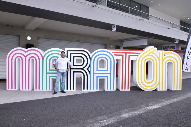 Ein Mann steht am Start eines Marathons vor einem großen "Marathon"-Schild, mit einem Banner rechts und einem Gebäude mit Lichtern, Geländern und Läden im Hintergrund.