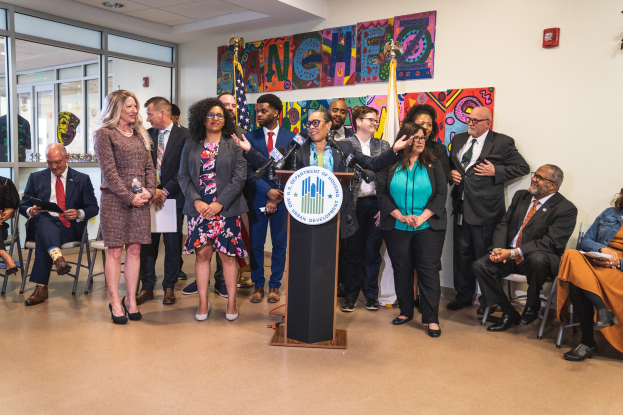 Gruppe von Menschen vor einem Rednerpult mit einem Mikrofon und einer Tafel mit der Aufschrift "Sancho Stadtrat tritt dem Stadtrat bei", mit sitzenden und stehenden Teilnehmern, Flaggen, einem Feuerlöscher und Deckenleuchten im Hintergrund.