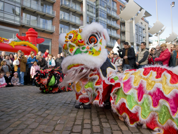 Vibrantes chinesisches Neujahrsfest in Amsterdam mit Löwen tanzen und einer Menge Schaulustiger, einige mit Kameras, vor einem Hintergrund aus Gebäuden, Laternenmasten und einem klaren blauen Himmel.