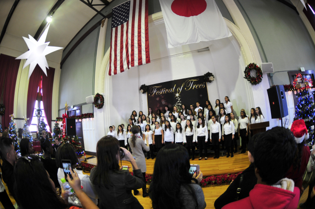 Eine Gruppe von Menschen auf einer Bühne vor einem Publikum, mit geschmückten Weihnachtsbäumen, einem Podium, Lautsprechern, einer Texttafel, Vorhängen, Flaggen und Deckenleuchten.