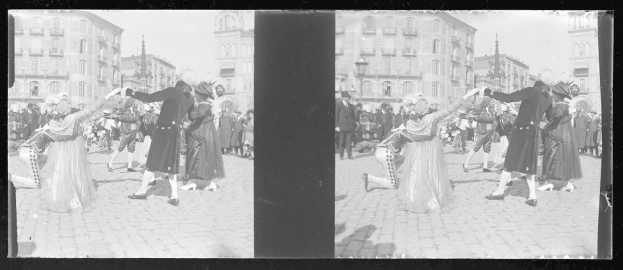 Ein Schwarz-Weiß-Foto einer Gruppe von Menschen, die auf der Straße tanzen, während eines Faschingsumzugs, mit Gebäuden, Straßenlaternen und Laternenpfählen im Hintergrund bei klarem Himmel.