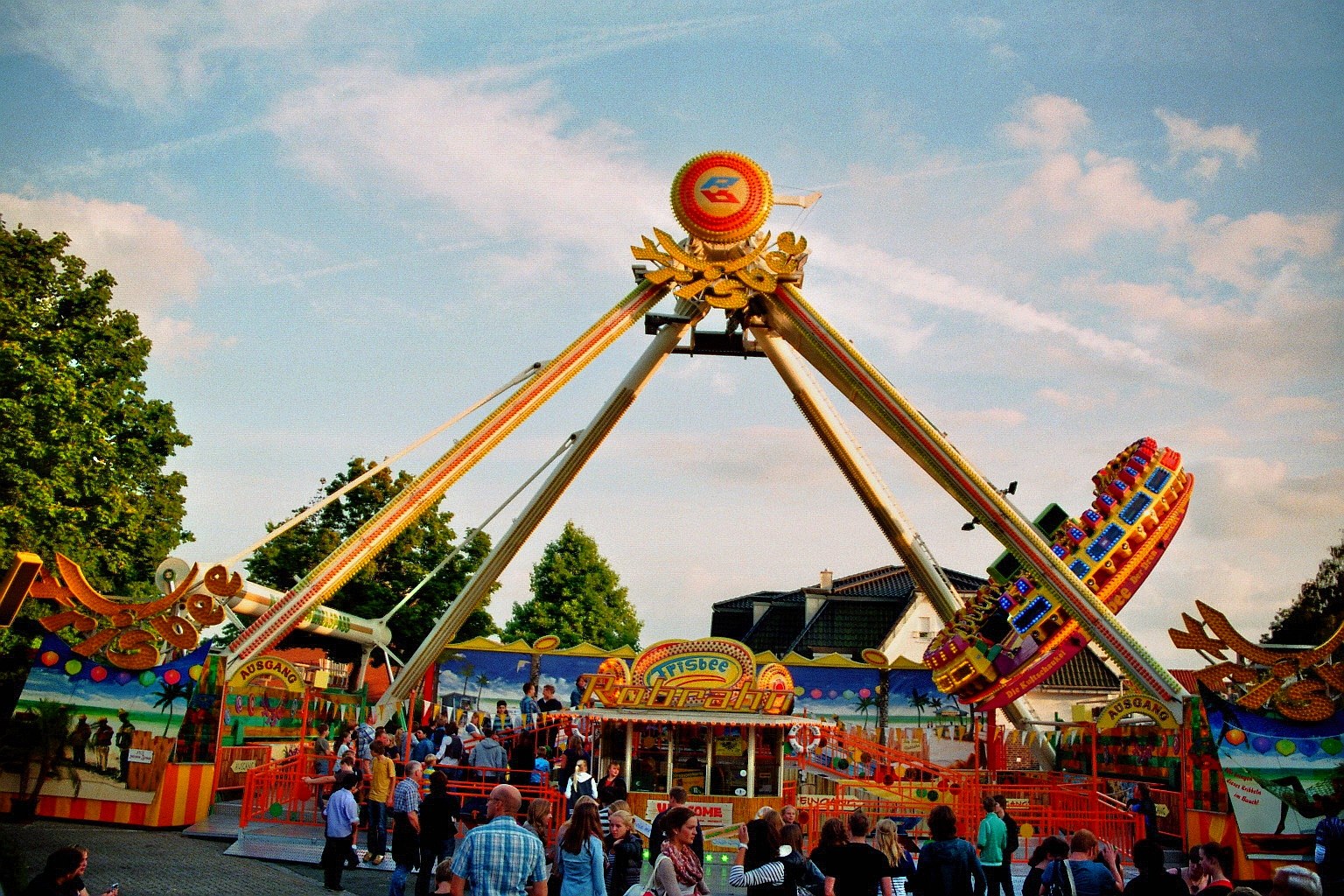 Eine Menschenmenge steht um eine Karussellfahrt auf einem Rummelplatz, umgeben von anderen Fahrgeschäften, Bäumen, Gebäuden und einem bewölkten Himmel.