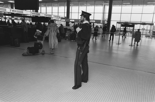 Ein Polizist in Uniform und Mütze steht in einem Flughafen-Terminal, hält eine Waffe und ist von Menschen und Gepäckwagen umgeben.