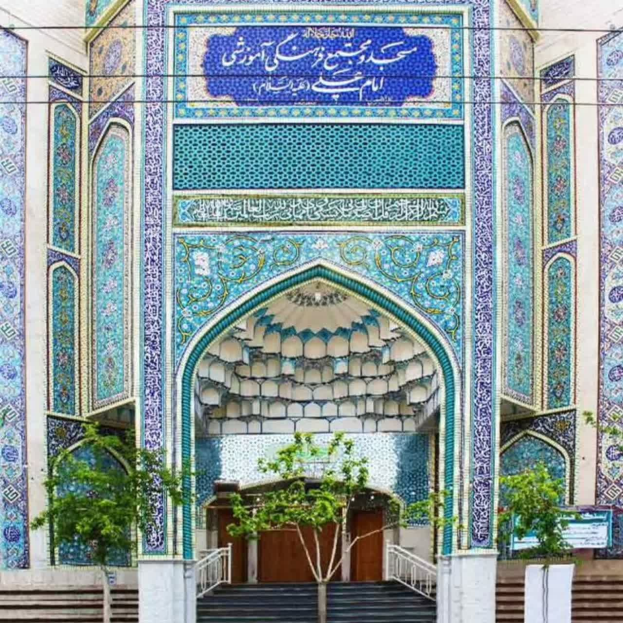 Blau-weiß gekachelte Eingangswand mit kunstvollen Designs und Text, Treppe hinauf zum Gebäude, Geländer auf beiden Seiten und Bäume im Vordergrund, mit einer Tafel mit Touristeninformationen über Isfahan, Iran.