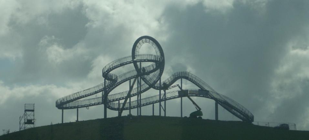 Eine Achterbahn auf dem Höhepunkt eines Hügels unter einem bewölkten Himmel, mit sichtbaren Stützpfählen und -strukturen.