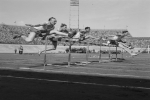Eine Gruppe von Männern in Schwarz-Weiß springt über Hürden auf einer Laufbahn, im Hintergrund ist ein Stadion mit Zuschauern und eine Turm in der Ferne zu sehen.