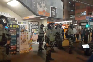 Eine Gruppe von Polizisten in Uniform steht vor einer Menge von Demonstranten mit Helmen und Waffen, mit Plakaten an den W├Ąnden, Lampen an der Decke und Geb├Ąuden mit Fenstern im Hintergrund, in Hong Kong.