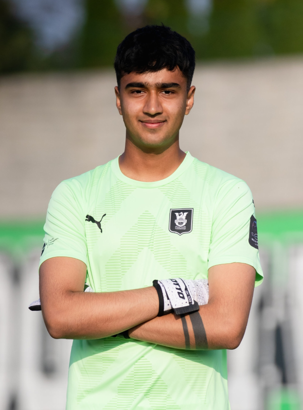 Ein junger Mann in einem grünen Hemd steht auf einem Fußballfeld mit verschränkten Armen und lächelt, mit einem unscharfen Hintergrund, der darauf hindeutet, dass er der Fokus des Bildes ist.