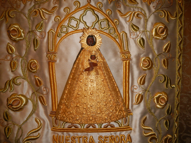 Goldverzierte Chasuble mit einer friedlichen Frau in einem hochgeschlossenen, langärmligen Kleid mit einem Dutt, umgeben von detaillierten Designs und prunkvollen Texten in einem goldenen Rahmen.