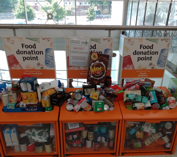 Orange Schränke gefüllt mit Lebensmittelspenden einschließlich Flaschen, Dosen und Kartons, mit Informationsschildern, vor einem Hintergrund aus Fahrzeugen, Bäumen, Gebäuden und Himmel durch Glasfenster sichtbar.