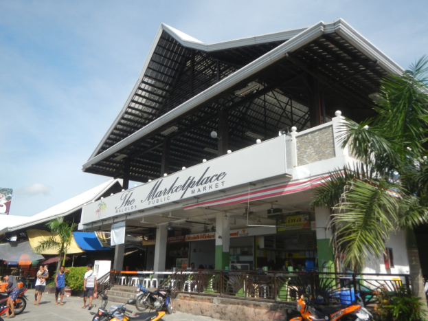 Marktplatz öffentlicher Markt in Koh Samui, Thailand, mit Menschen, geparkten Fahrzeugen, einem Metallzaun, Bäumen, einem Banner, Pfosten, einem Dach mit Deckenlampen und einem bewölktem Himmel.