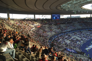Große Menschenmenge in einem Stadion bei einem Fußballspiel, mit einer Bühne, Fahnen, Stangen, einem Bildschirm und der Allianz Arena in München, Deutschland im Hintergrund.