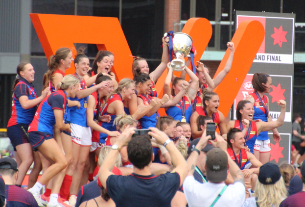 Gruppe von Frauen-AFL-Spielerinnen feiert mit einem Pokal vor einer jubelnden Menge, einige halten den Pokal hoch oder fotografieren den Moment mit Geräten, mit Anzeigetafeln und einem Glasgebäude im Hintergrund.