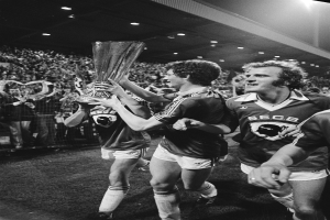 Eine Gruppe von Männern hält einen Pokal auf einem Fußballfeld hoch, feiert ihren Sieg im FA-Cup-Finale, umgeben von einer Menge hinter einem Zaun, unter Stadionbeleuchtung, in Schwarz-Weiß.