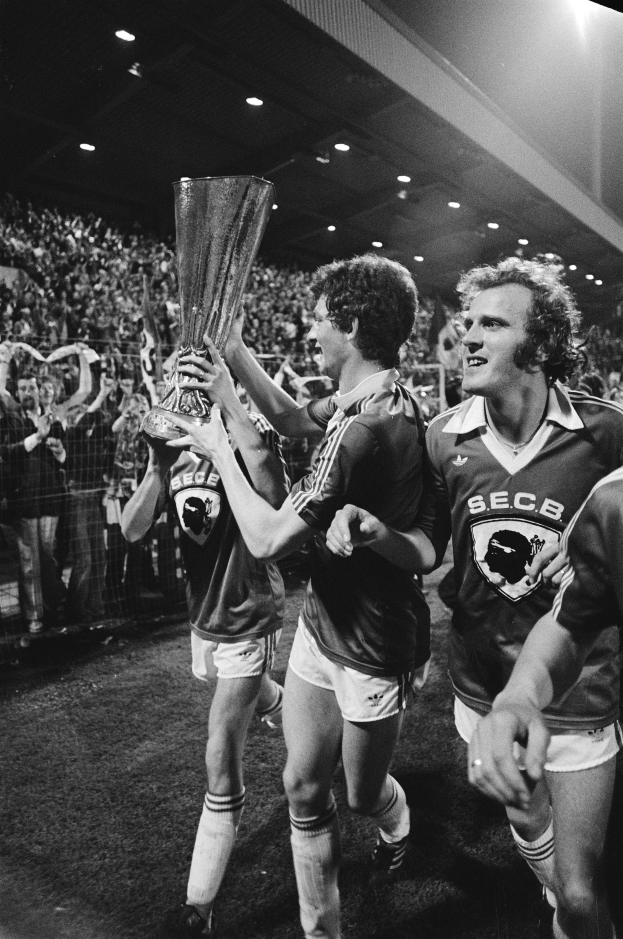 Eine Gruppe von Männern hält einen Pokal auf einem Fußballfeld hoch, feiert ihren Sieg im FA-Cup-Finale, umgeben von einer Menge hinter einem Zaun, unter Stadionbeleuchtung, in Schwarz-Weiß.