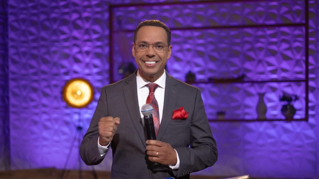 Stephen Colbert, Moderator von *The Daily Show with Trevor Noah*, steht auf einer Bühne und hält ein Mikrofon in der Hand, trägt einen Anzug, Krawatte und eine Brille, während hinter ihm Lichtausrüstung und Gegenstände zu sehen sind.