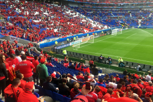 Ein volles Stadion mit Sitz- und Stehplätzen, Fahnen, einem Tor und Deckenleuchten, während die Menge ungeduldig auf den Beginn eines Fußballspiels wartet.