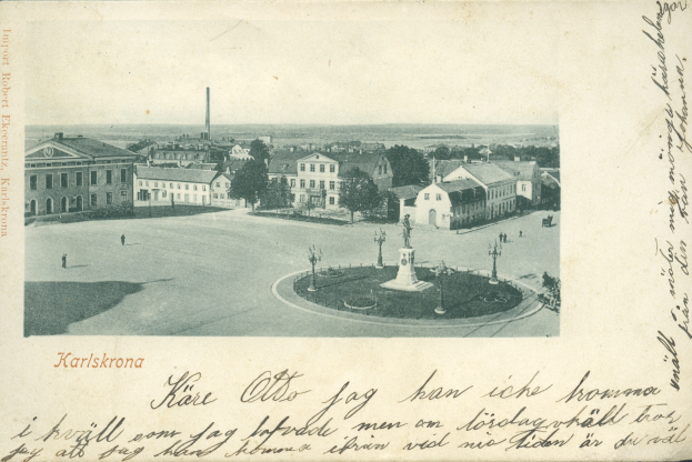 Eine alte Postkarte zeigt eine Stadtansicht mit Gebäuden, Bäumen, Masten und einem zentralen Brunnen, mit handgeschriebener Schrift am unteren Rand.