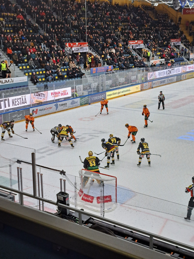 Eine Gruppe von Menschen, die Hockey auf einem Eisring in einer Arena spielen, mit einem Netz, einer Umzäunung, sitzenden Zuschauern, Bannern und Scheinwerfern.