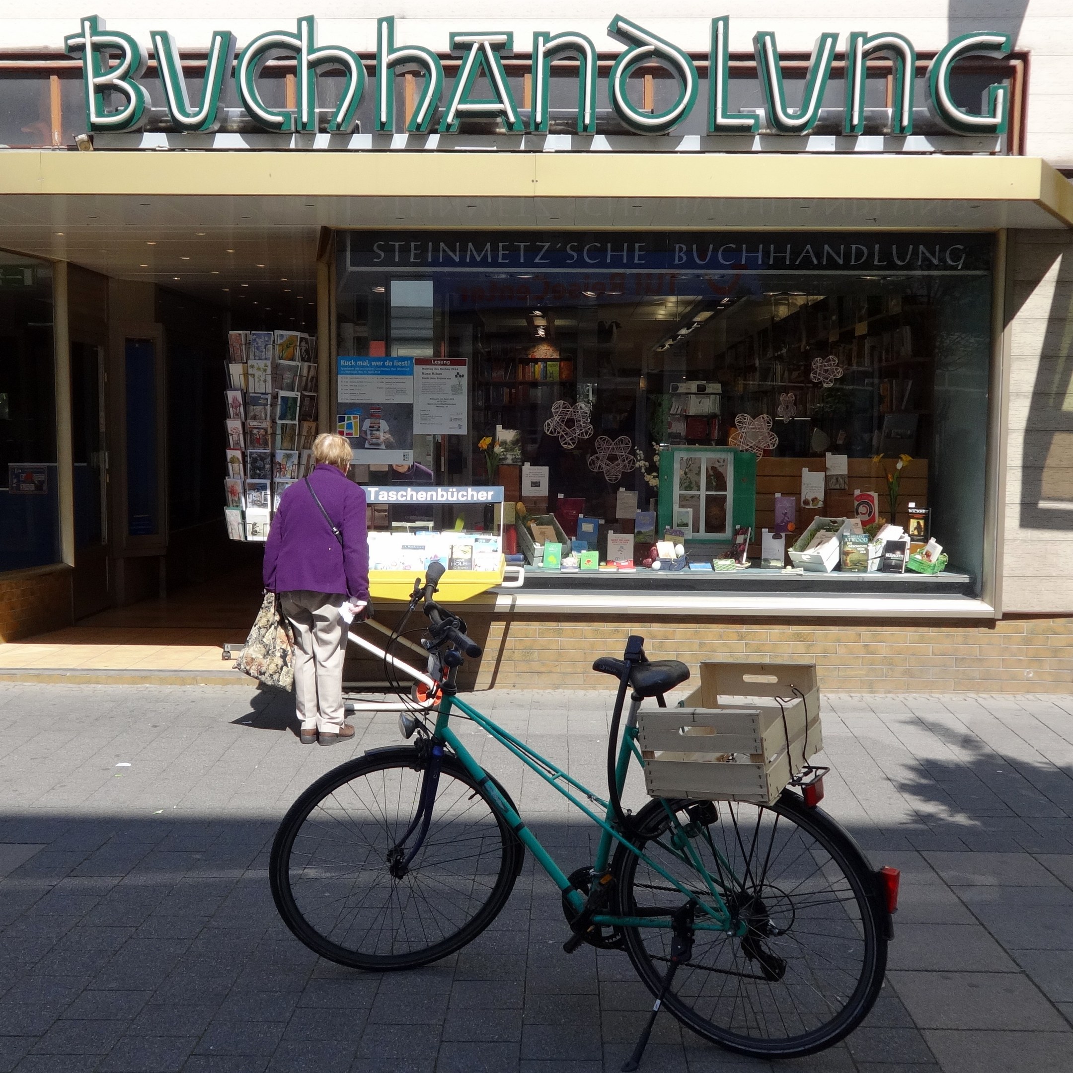 Eine Frau steht neben einem Fahrrad, das vor einem Laden geparkt ist, mit einem Fahrradrad in Vordergrund und Büchern und anderen Gegenständen durch die Gläserscheibe des Ladens sichtbar.