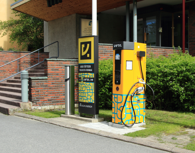 Eine Elektrofahrzeug-Ladestation an der Straße, umgeben von Grün und einem Gebäude mit Fenstern und Säulen, mit Treppen und Geländern, die zu ihr führen.