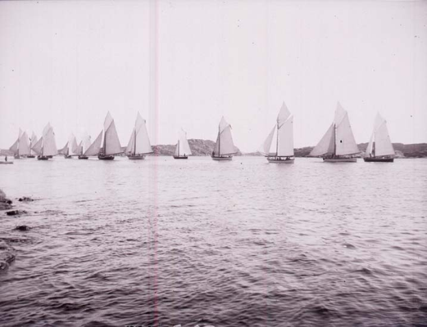 Schwarzes und weißes Foto einer Gruppe von Segelbooten auf dem Wasser mit Hügeln im Hintergrund und einem klaren Himmel, beschriftet als Foto des New York Yacht Club.
