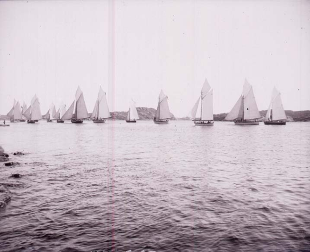 Schwarzes und weißes Foto einer Gruppe von Segelbooten auf dem Wasser mit Hügeln im Hintergrund und einem klaren Himmel, beschriftet als Foto des New York Yacht Club.