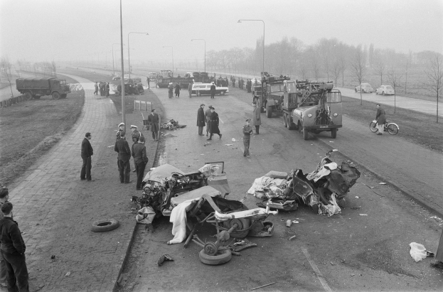 Schwarz-weiß-Szene eines Autounfalls am Straßenrand mit mehreren Fahrzeugen, Einsatzkräften und Schaulustigen in der Nähe des beschädigten Autos, umgeben von Laternen, Bäumen und Himmel.