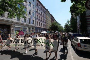 Eine Gruppe von Menschen marschiert auf der Straße in Berlin, hölt eine Fahne, mit geparkten Autos auf der rechten Seite, einem Radfahrer auf der linken Seite und Gebäuden, Bäumen und einer Schautafel im Hintergrund unter einem sichtbaren Himmel.