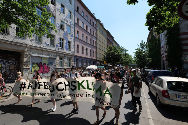 Eine Gruppe von Menschen marschiert auf der Straße in Berlin, hölt eine Fahne, mit geparkten Autos auf der rechten Seite, einem Radfahrer auf der linken Seite und Gebäuden, Bäumen und einer Schautafel im Hintergrund unter einem sichtbaren Himmel.