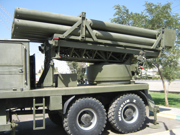 Militärlastwagen mit einer Rakete auf der Rückseite, der auf einer Straße fährt, umgeben von Gras, Bäumen, Gebäuden und einem Maschendrahtzaun.