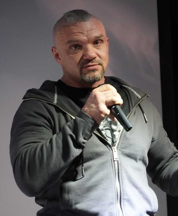 Ein Mann in einer schwarzen Kapuzenjacke und einem schwarzen Anzug mit einem weißen Hemd hält ein Mikrofon und steht selbstbewusst vor einer Wand, wobei er wie ein Comedian wirkt.