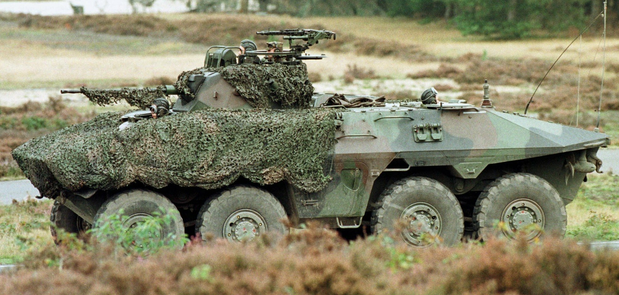 Militärischer Mannschaftstransporter mit einer montierten großen Kanone, der auf einer Straße neben einem Wald fährt.