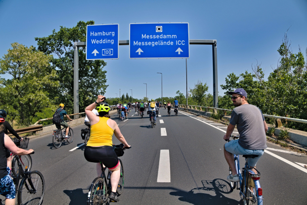 Gruppe von Radfahrern mit Helmen auf einer Straße mit einer Begrenzung auf einer Seite und Bäumen auf der anderen unter einem klaren blauen Himmel mit Laternen im Hintergrund und einer Tafel, die eine Radtour in Hamburg anzeigt.