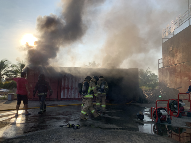 Eine Gruppe von Feuerwehrleuten in Helmen steht vor einem brennenden Gebäude mit aufsteigendem Rauch, während auf der rechten Seite ein Gebäude mit einer Namenschilder und links Bäume zu sehen sind.