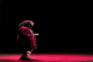 A woman stands on a red carpet in a dark room, wearing a dress and footwear, ready for a performance.