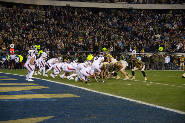 Ein Fuballspiel in einem Stadion mit Spielern in Sportkleidung und Helmen auf dem Feld, Zuschauern, Gelöndern und Tafeln mit Text im Hintergrund.